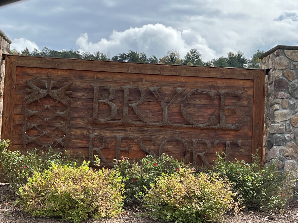 A wooden sign displays the name 'Bryce Resort,' framed by a stone structure. Lush green shrubs and ornamental plants surround the base, complemented by a backdrop of trees under a partly cloudy sky.