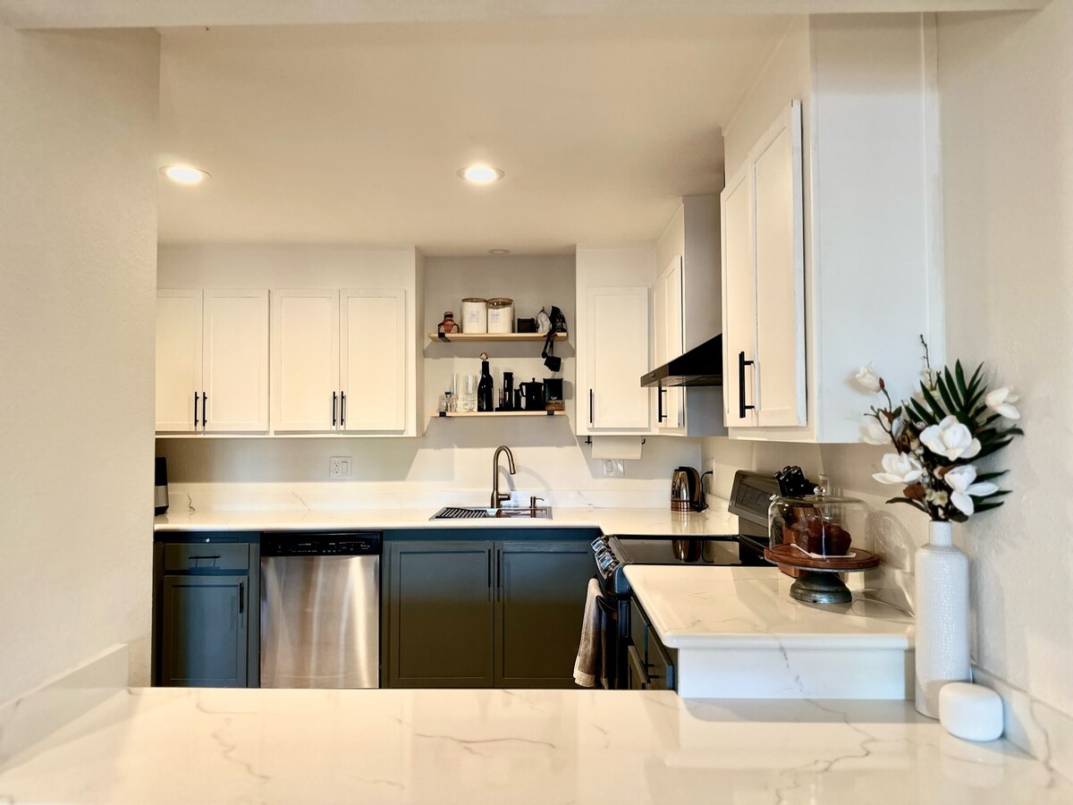 A modern kitchen is presented with white cabinetry and dark lower cabinets, complemented by a marble countertop. The kitchen includes a stainless steel dishwasher, a sink, and a stove. Shelves display kitchen essentials, while a small vase with flowers adds a touch of decor.