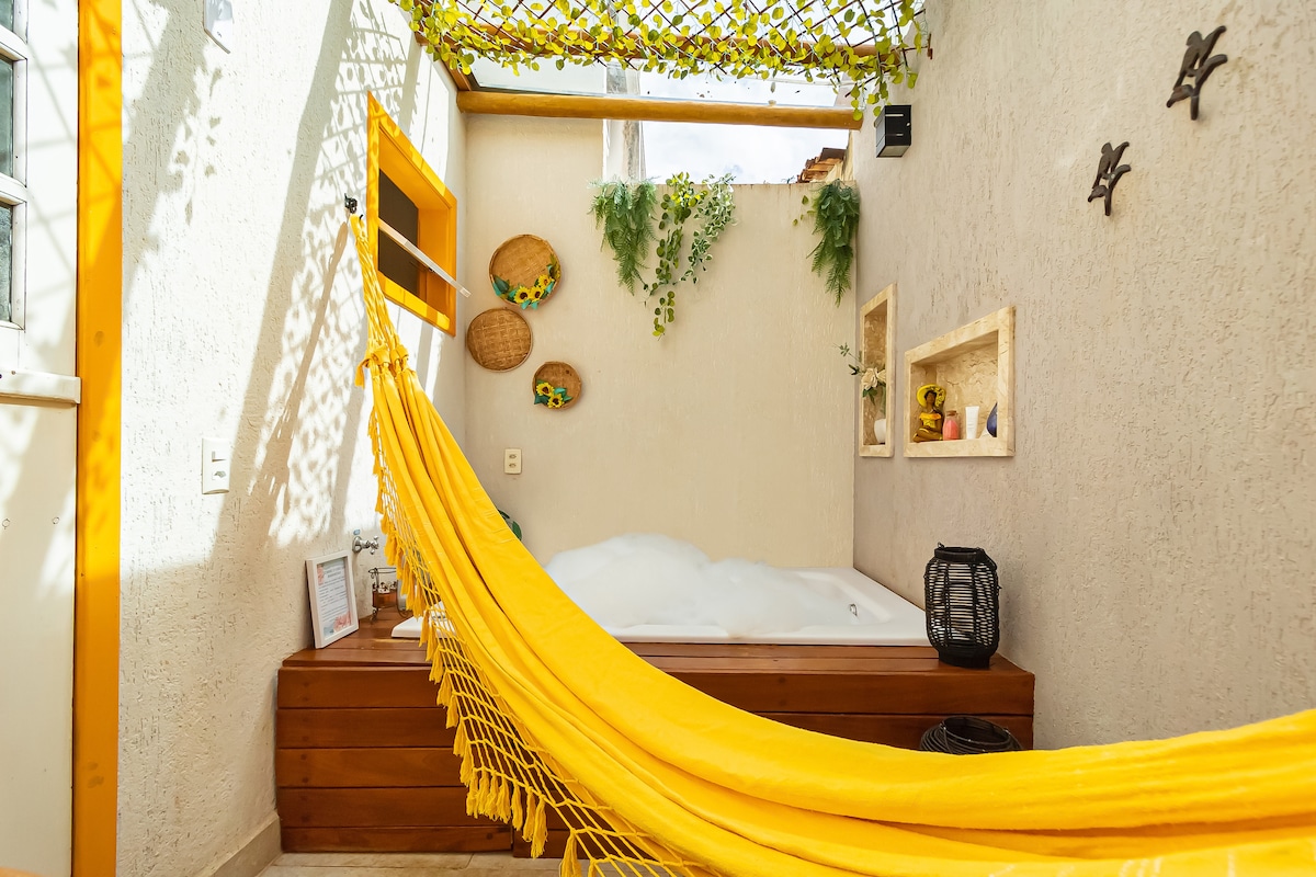 A private area features a double hot tub surrounded by wooden accents, with a vibrant yellow hammock draped nearby. The walls are adorned with green plants and decorative items, creating a relaxing environment infused with natural light from an overhead lattice.