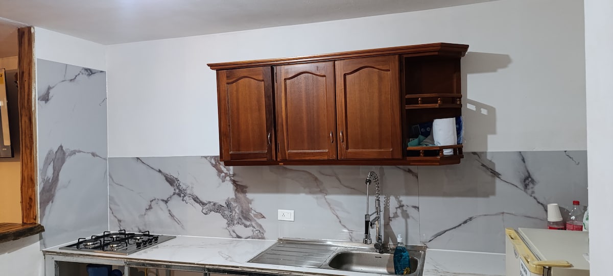 A kitchen area is presented with wooden cabinetry above a marble-patterned backsplash. A stainless steel sink is fitted into the countertop, complemented by a gas stove. Light fixtures are installed in the ceiling, ensuring a well-lit cooking space.