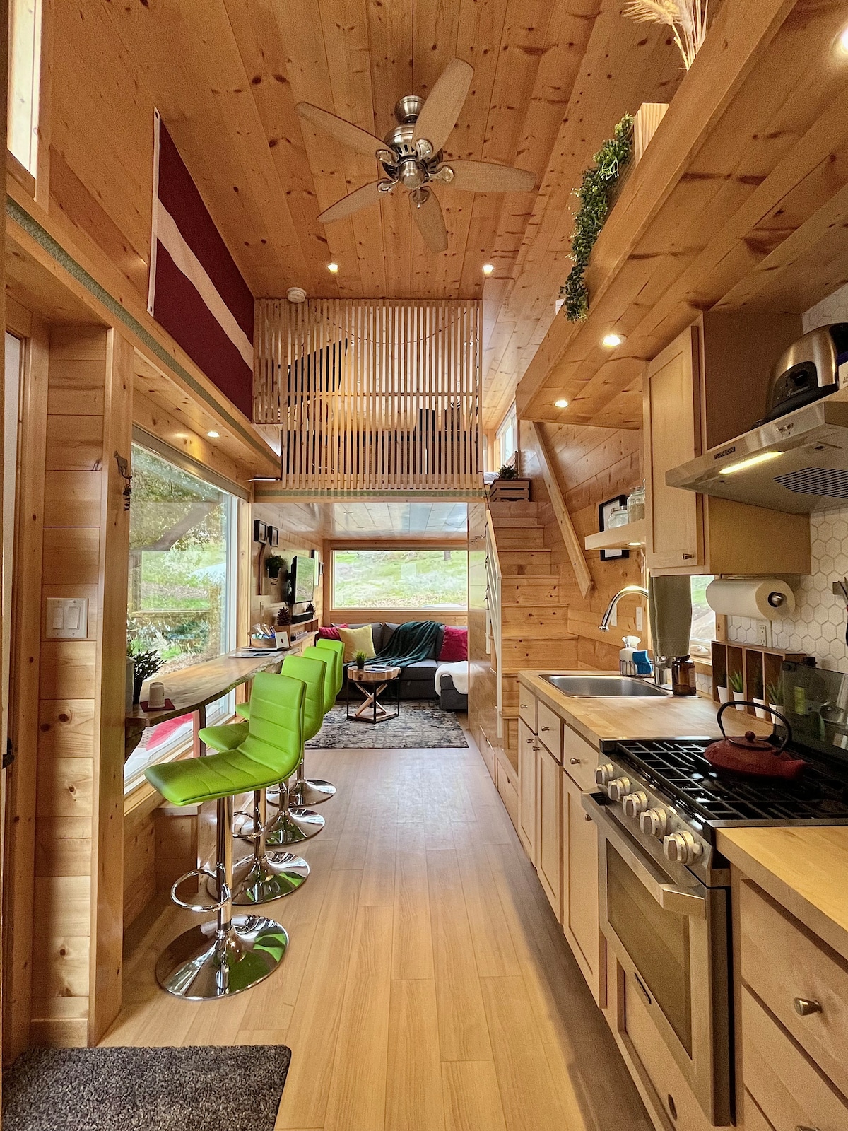A narrow corridor is highlighted by natural wood paneling, leading to a cozy living area and a staircase. Barstools with green seats line a counter near the window, while kitchen appliances and a gas range provide essential cooking amenities.