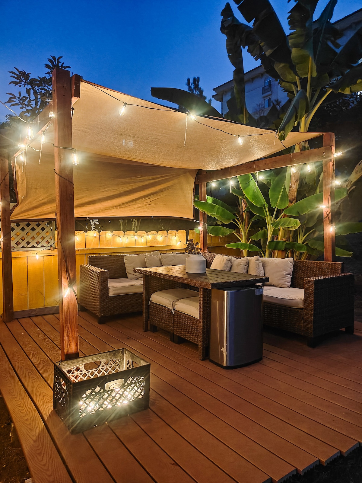 An inviting outdoor sitting area features comfortable wicker furniture, complemented by soft cushions. A shaded pergola is adorned with string lights, creating a cozy ambiance. Lush greenery, including banana plants, surrounds the space, enhancing the natural setting. A small fridge and table are accessible for convenience.
