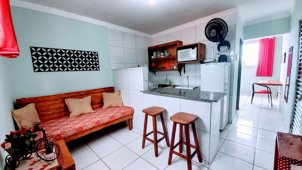 A cozy kitchen area features a granite countertop and a microwave, with bar stools positioned nearby. A wooden bench with decorative cushions offers seating, complemented by a small dining table. Bright red curtains frame the windows, adding a pop of color to the space.