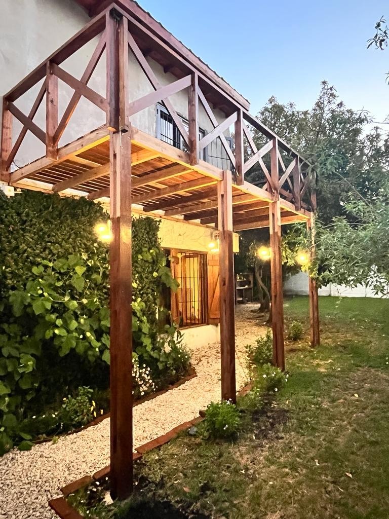 A charming exterior view showcases a wooden balcony elevated over a pebbled pathway. Lush greenery adorns the wall, while softly glowing lanterns provide ambient lighting along the path. The surrounding garden features well-maintained grass and plants, contributing to a serene atmosphere.