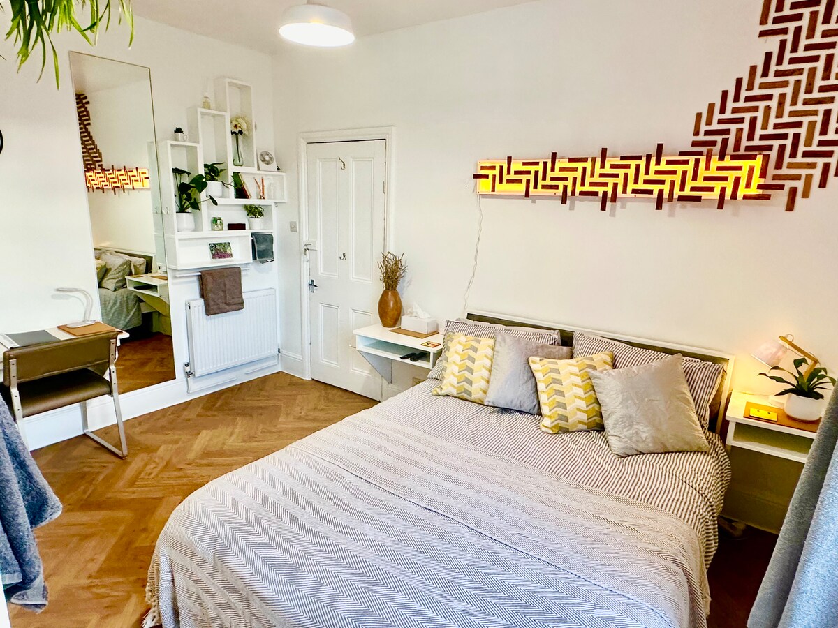 A spacious bedroom features a comfortable bed adorned with patterned pillows. Wall-mounted light fixtures provide soft illumination, while stylish shelving holds plants and decor. A mirror reflects the room's ample natural light, and a minimal dining space is visible, highlighting a harmonious design.