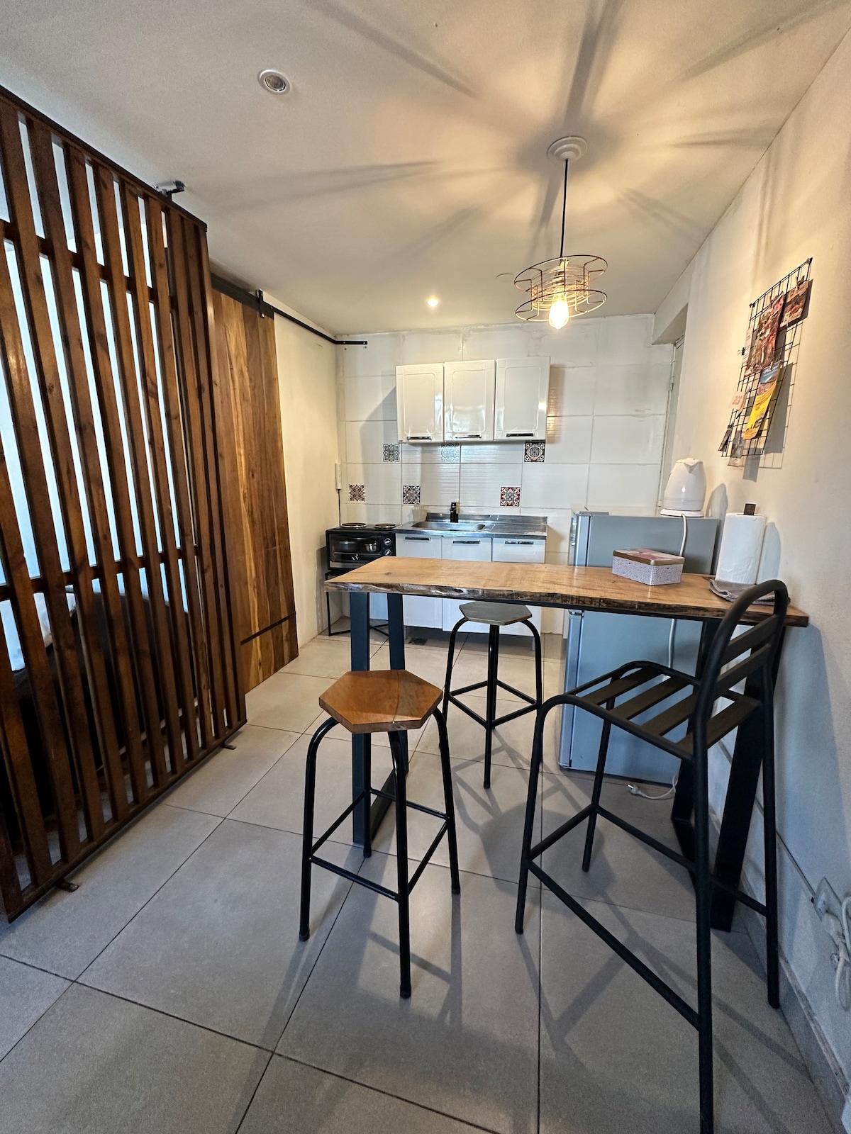 A modern kitchen area is visible, featuring sleek cabinetry and integrated appliances. A wooden divider adds warmth to the space. There is a small dining table with two stools, enhancing the functional layout. Soft lighting illuminates the room, contributing to a bright and inviting atmosphere.