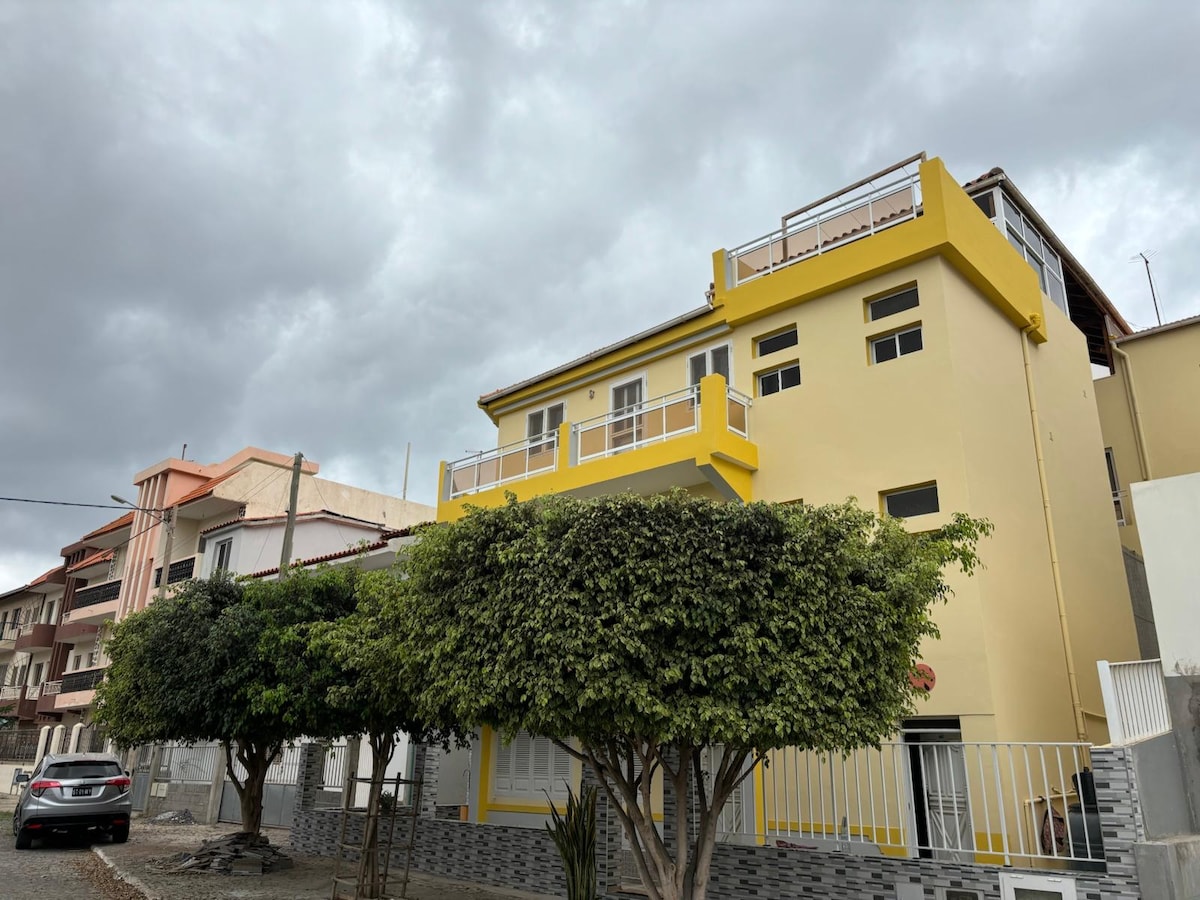 A two-story, yellow building is surrounded by lush green trees. A spacious rooftop terrace is visible, along with large windows that illuminate the structure. The neighborhood features residential buildings and a clear sky with clouds.