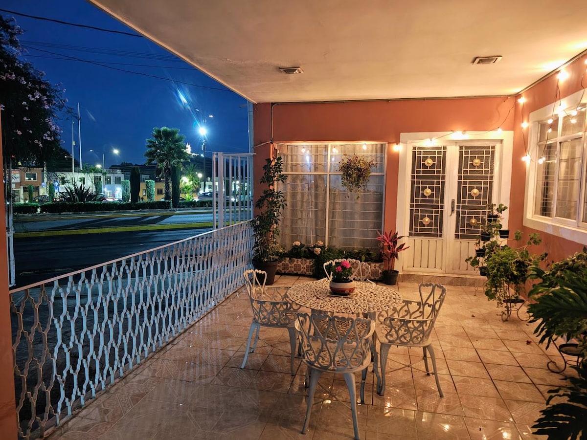 A covered outdoor terrace is presented, featuring a round table surrounded by white metal chairs. Ambient lights illuminate the space, enhancing the greenery with potted plants nearby. The entrance is framed by large windows, offering a view of the surroundings.
