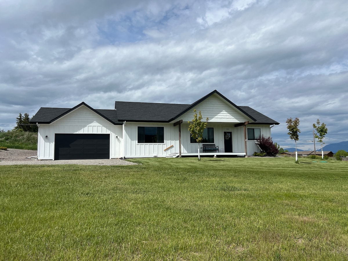 A ranch-style home is depicted with a white exterior and a dark roof. The front yard features a well-maintained lawn and a gravel driveway leading to a finished garage. Scenic mountain views and a cloudy sky are visible in the background.