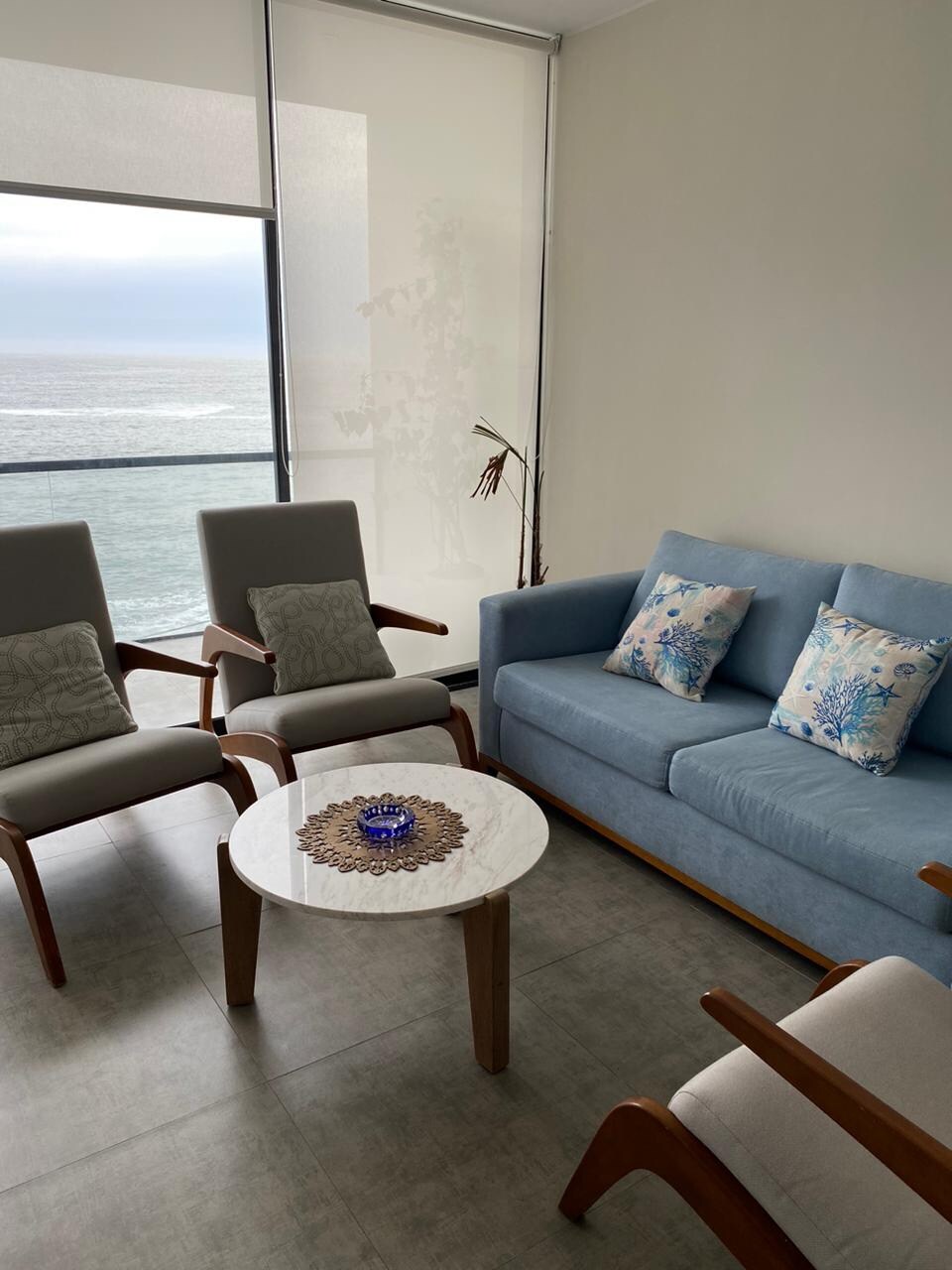 A bright seating area is presented with a light blue sofa and two wooden chairs. Decorative cushions add a touch of color. A small round table with a marble top sits centrally, offering a view of the ocean through large glass windows that invite natural light.