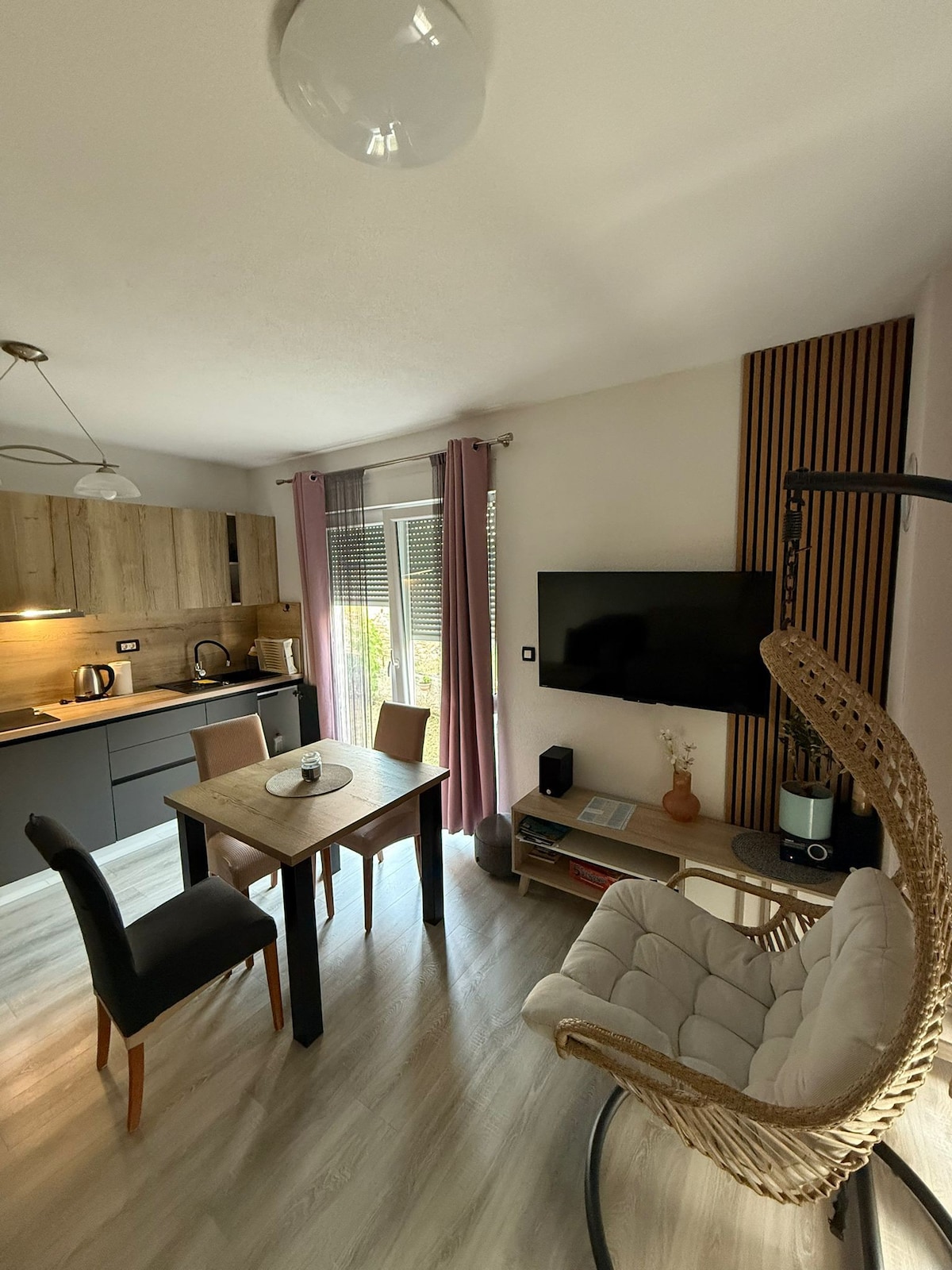 An inviting living space features a cozy dining table with four chairs, a comfortable hanging chair, and a television mounted on a wooden accent wall. Natural light pours in through large windows, enhancing the modern kitchen area equipped with essential appliances.