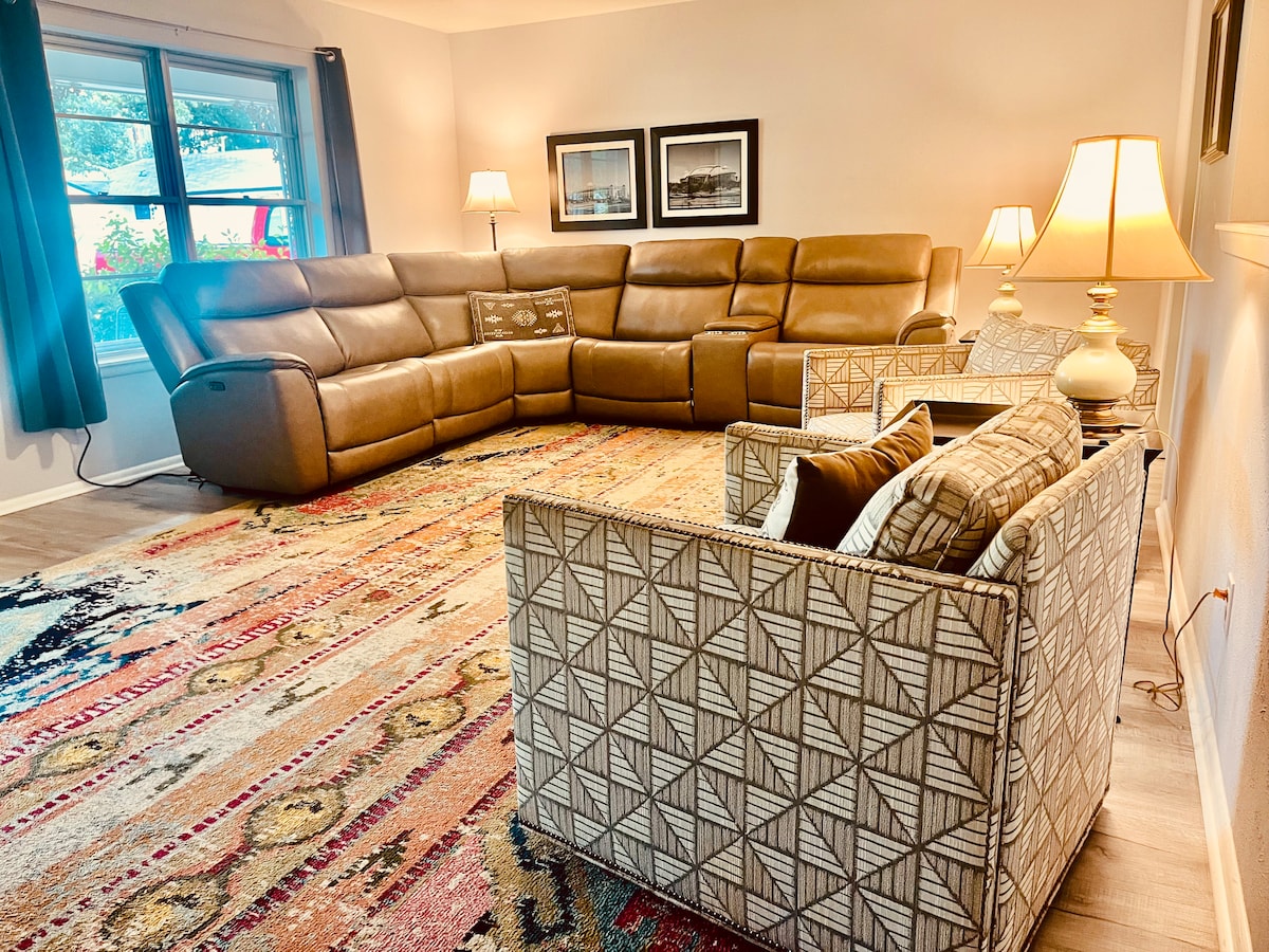 A cozy living area is presented with a large sectional sofa and an accent chair. Warm lighting is provided by table lamps, while natural light enters through two windows with dark curtains. The space is unified by a colorful area rug featuring diverse patterns.