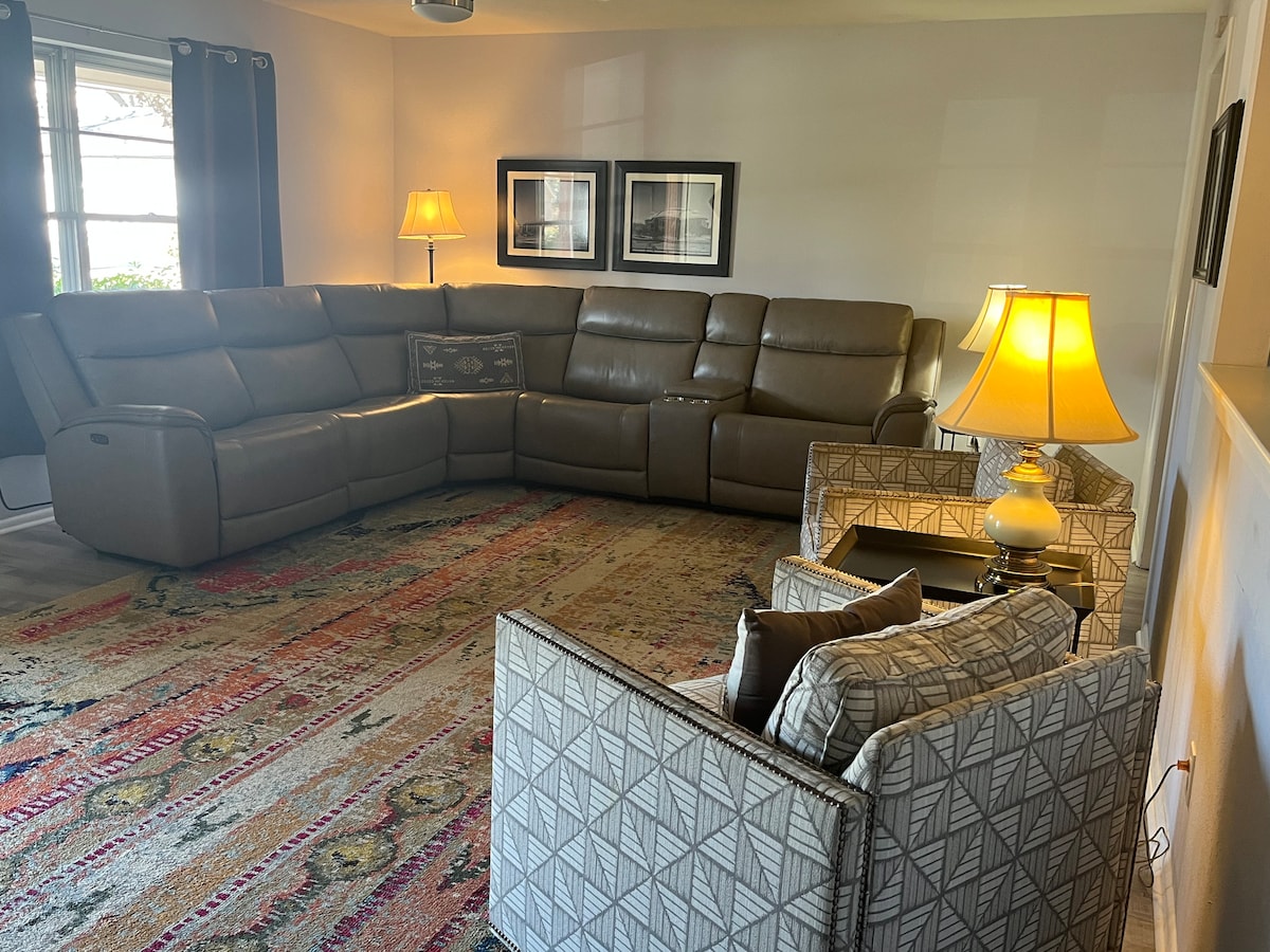 A comfortable living area features a large sectional sofa in a neutral tone and two patterned chairs. Soft lighting is provided by a floor lamp and a table lamp, creating a cozy atmosphere. A colorful area rug with intricate patterns adds warmth to the space.