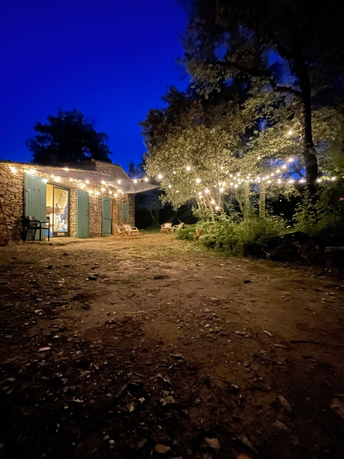 The outdoor area is softly illuminated by string lights, creating a warm ambiance against the night sky. A stone structure can be seen in the background, with open doors revealing a glimpse of the interior. Surrounded by trees and greenery, the space offers a tranquil setting.
