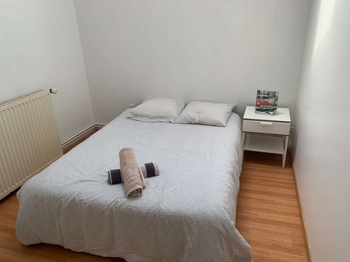 A cozy bedroom features a spacious bed with light bedding, complemented by two pillows. A nightstand with a decorative item is positioned next to the bed. Natural light brightens the room, and a radiator is visible on the wall.