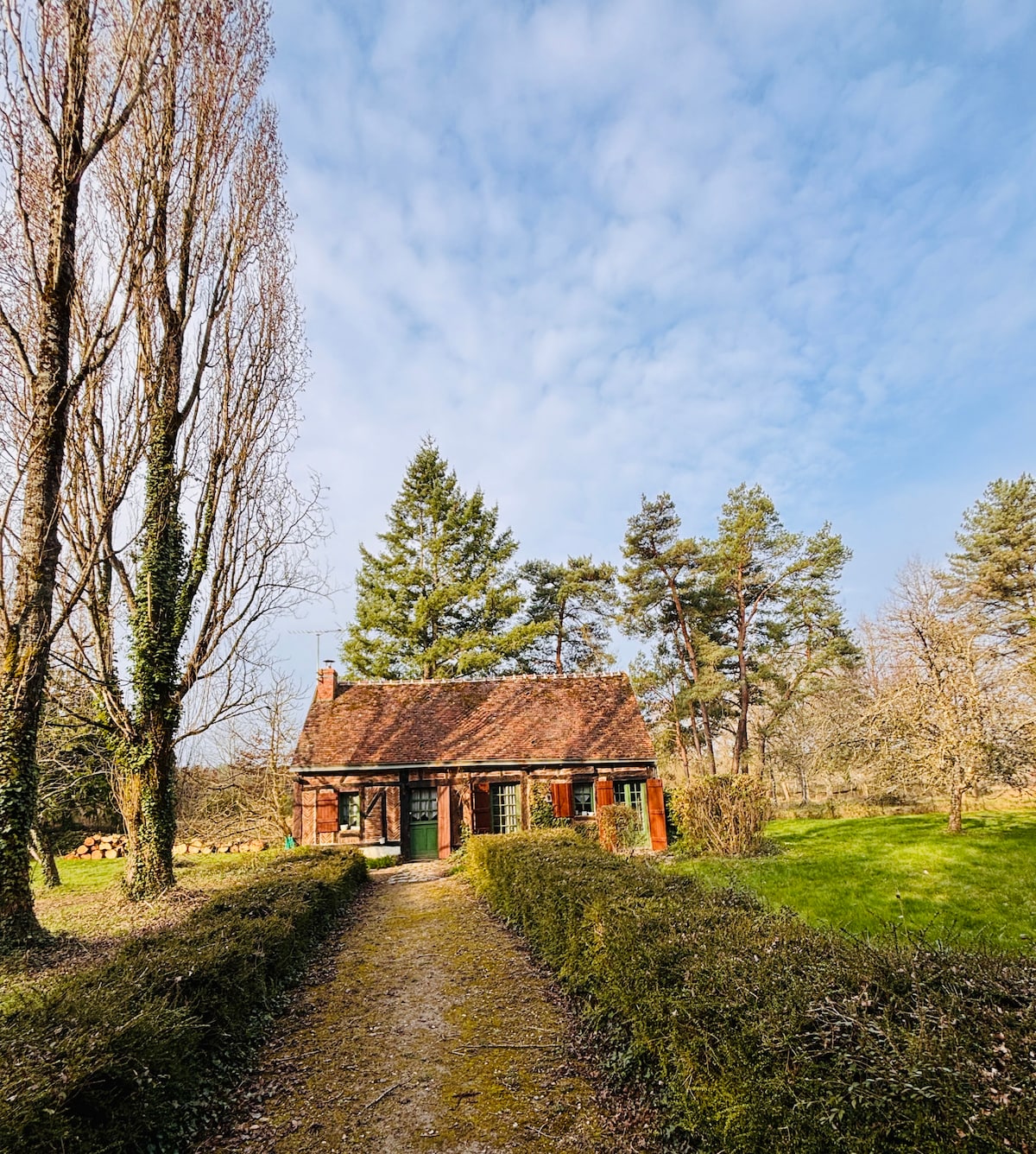 A charming house is positioned along a well-maintained pathway, surrounded by lush greenery and tall trees. Sunlight filters through the clouds, illuminating the rustic exterior with a gabled roof and large windows, while a manicured hedge lines the sides of the path.