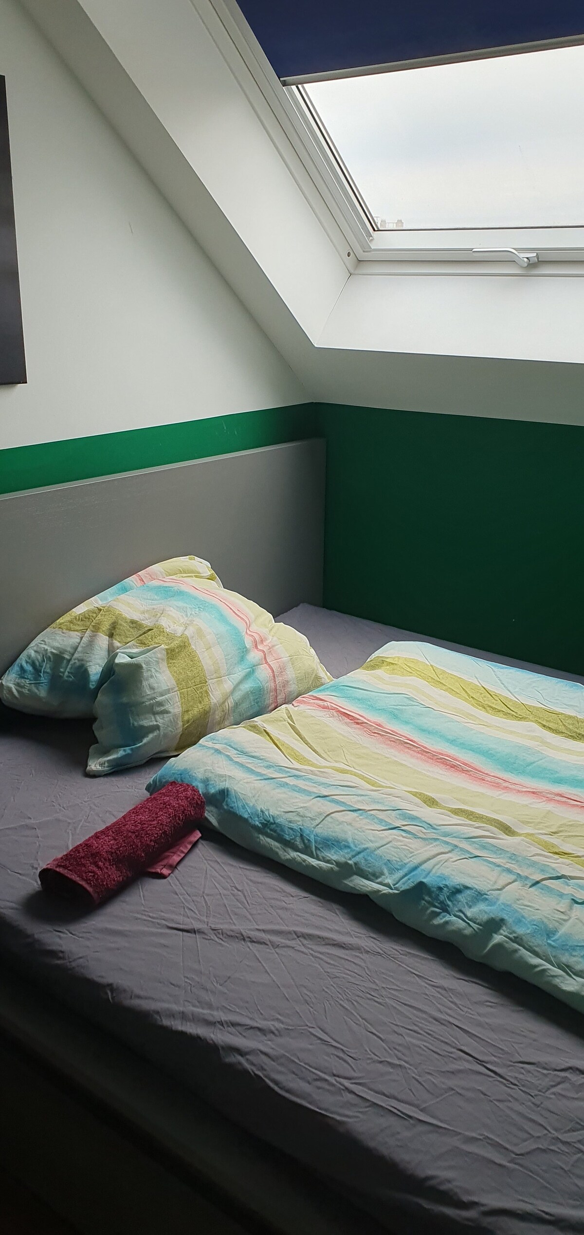 A cozy sleeping space features a compact bed with a striped duvet and two pillows. A towel is neatly placed on the blanket, and sunlight filters through a skylight above. Green accents on the walls add a touch of color.