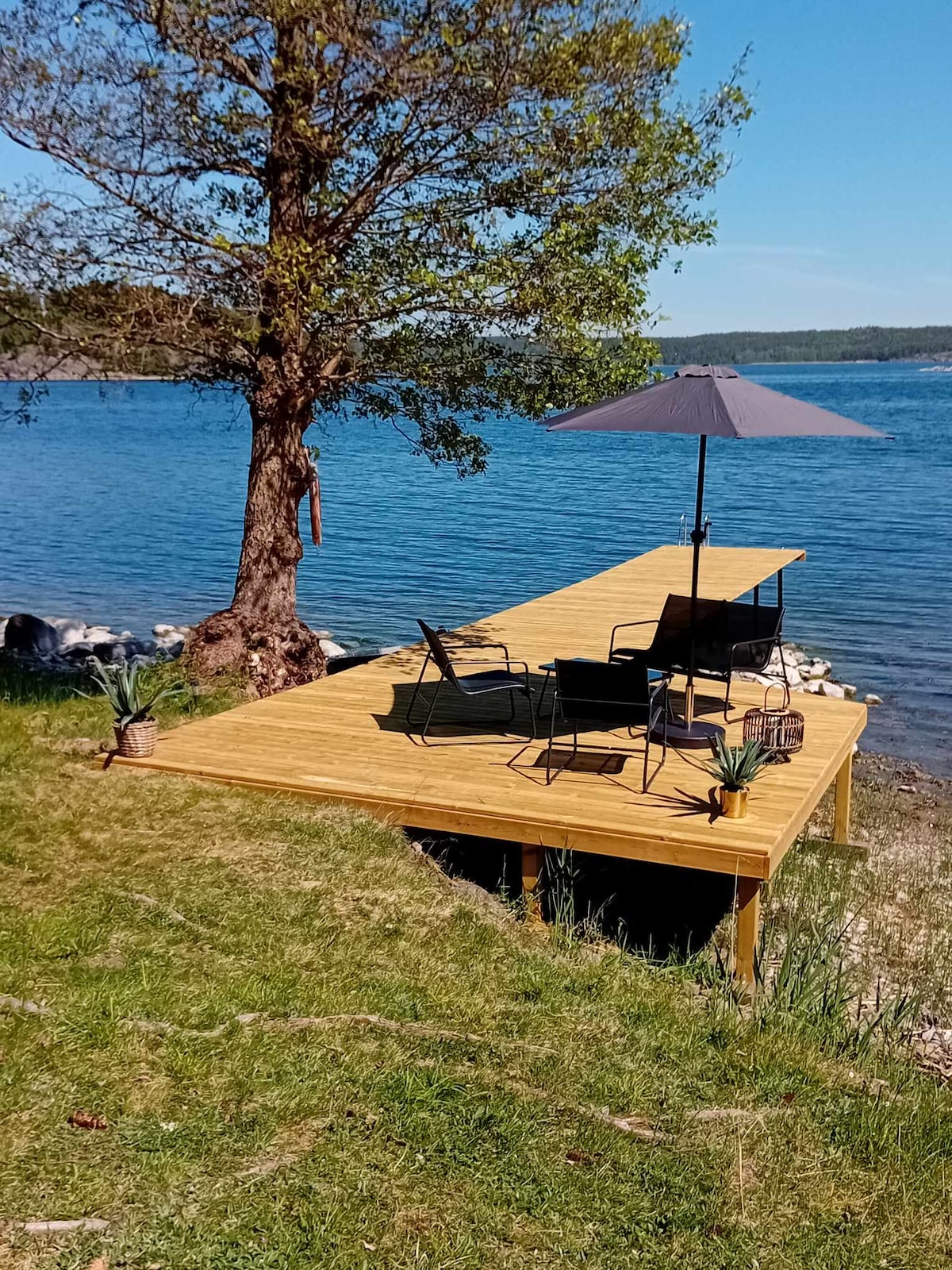 A sunny wooden dock extends toward tranquil waters, featuring two black chairs and a small table beneath a gray umbrella. A tree offers shade, while the grassy area enhances the serene waterfront setting. The calm water reflects the clear blue sky, creating a peaceful environment.