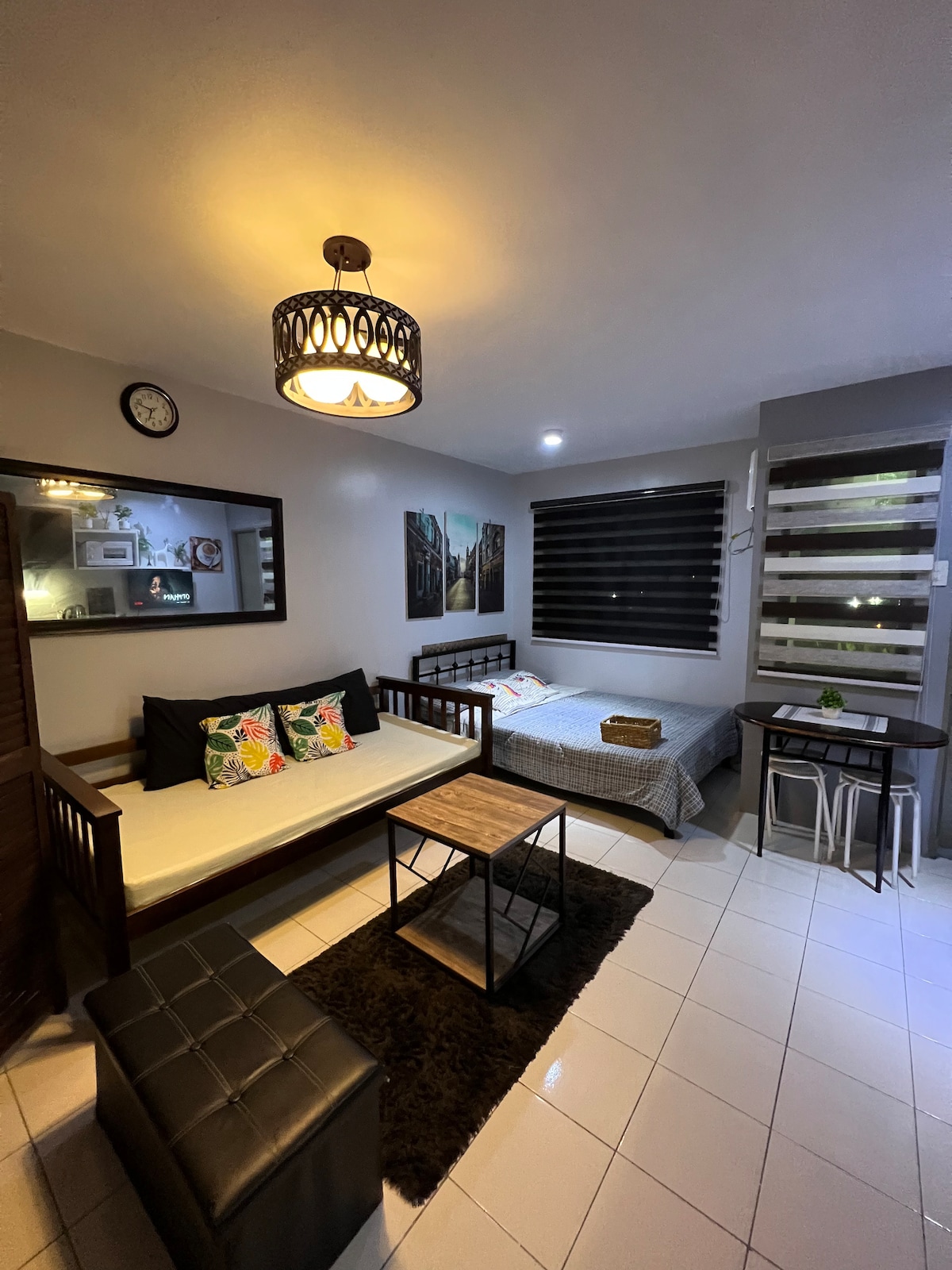 A cozy living area is presented with a daybed adorned with decorative pillows and a pull-out bed beside it. A coffee table sits on a soft rug, while a mirror reflects the space. Large windows with dark blinds allow for controlled lighting in the room.