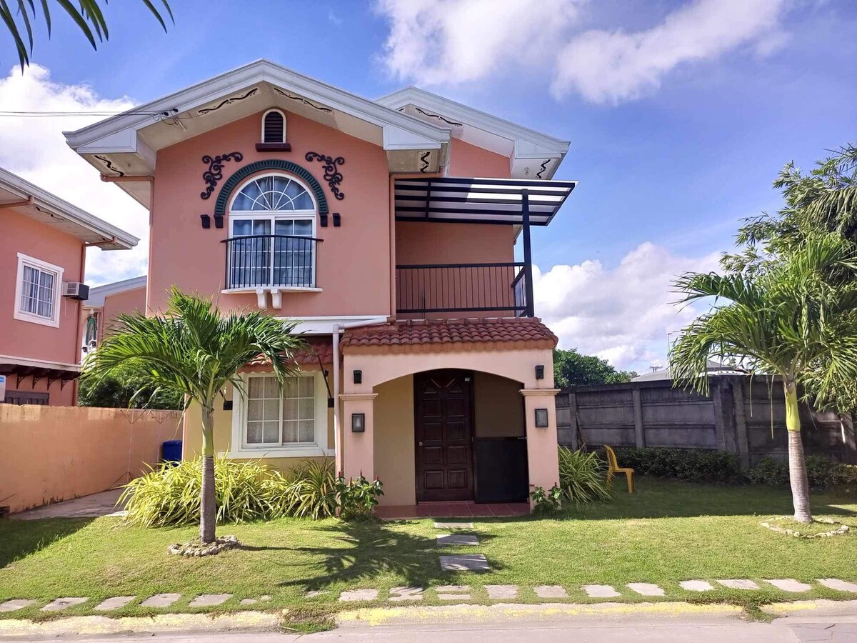 The exterior of the two-story house features a blend of pink and cream walls, adorned with decorative elements. A covered balcony extends from the upper level, while a lush green lawn surrounds the entrance. Tropical plants and a yellow chair enhance the outdoor space.