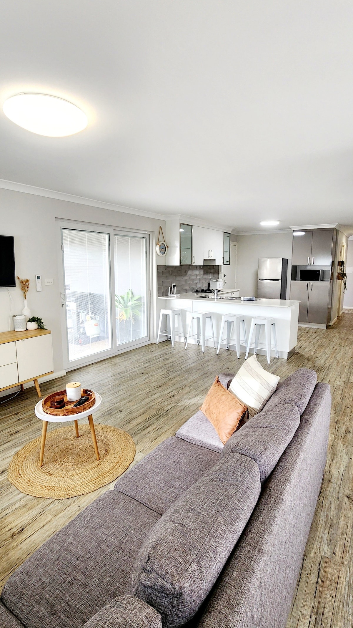 A modern living space is presented with a gray sofa and a small round table. A well-equipped kitchen with white cabinets and bar stools is visible in the background. Light streams through sliding glass doors, illuminating the neutral-toned decor and wooden flooring.