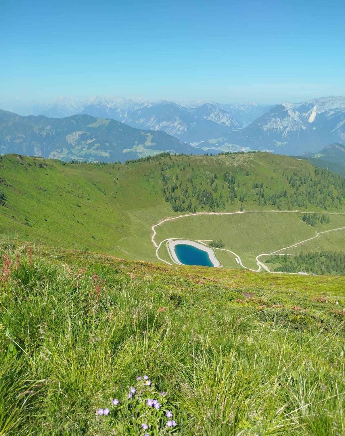 A panoramic view of a mountain landscape features rolling green hills adorned with wildflowers. A serene blue lake is visible below, flanked by winding paths. In the distance, majestic mountains rise under a clear blue sky.