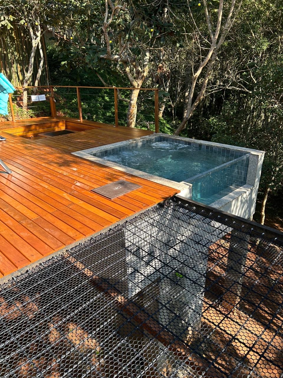 A wooden deck surrounds a jacuzzi, nestled within a natural setting. The jacuzzi is filled with water, and greenery is visible in the background, complementing the outdoorsy atmosphere. The deck features railings with a modern design, emphasizing the connection with nature.