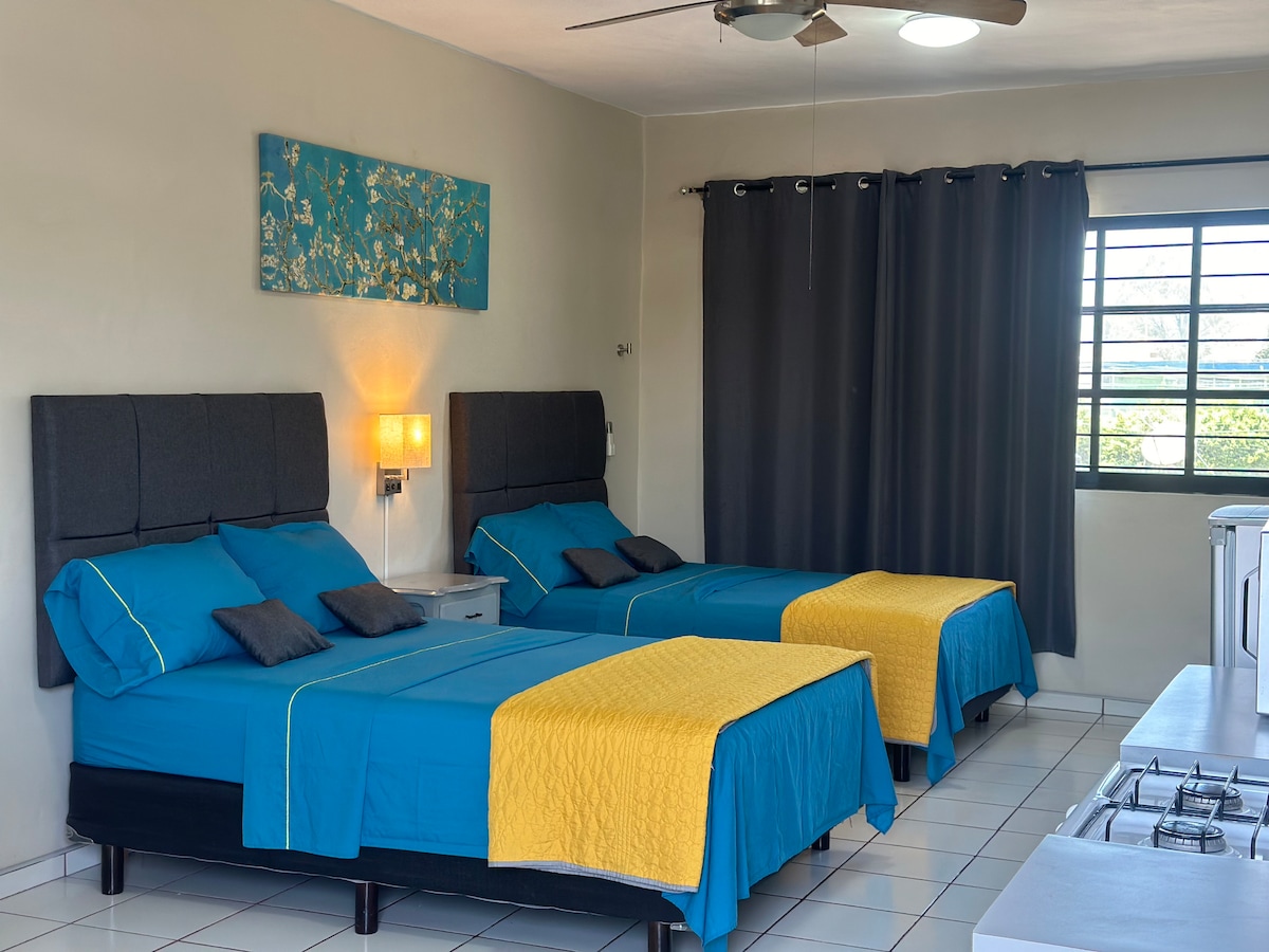 A well-lit room contains two spacious beds dressed in bright blue linens, accented with yellow throws. The neutral walls are complemented by a decorative wall piece above the beds. Large windows allow natural light, and dark curtains frame the view.