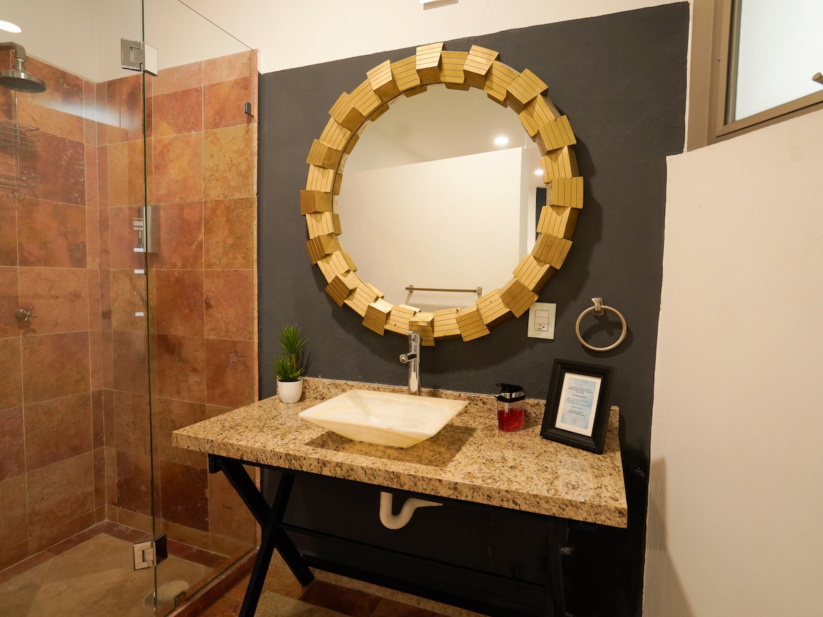 A modern bathroom showcases a stylish circular mirror framed in wood. A granite countertop holds a minimalist sink and decorative items, including a small plant. The shower area features a glass enclosure with tiled walls, complemented by warm, earthy tones in the overall design.