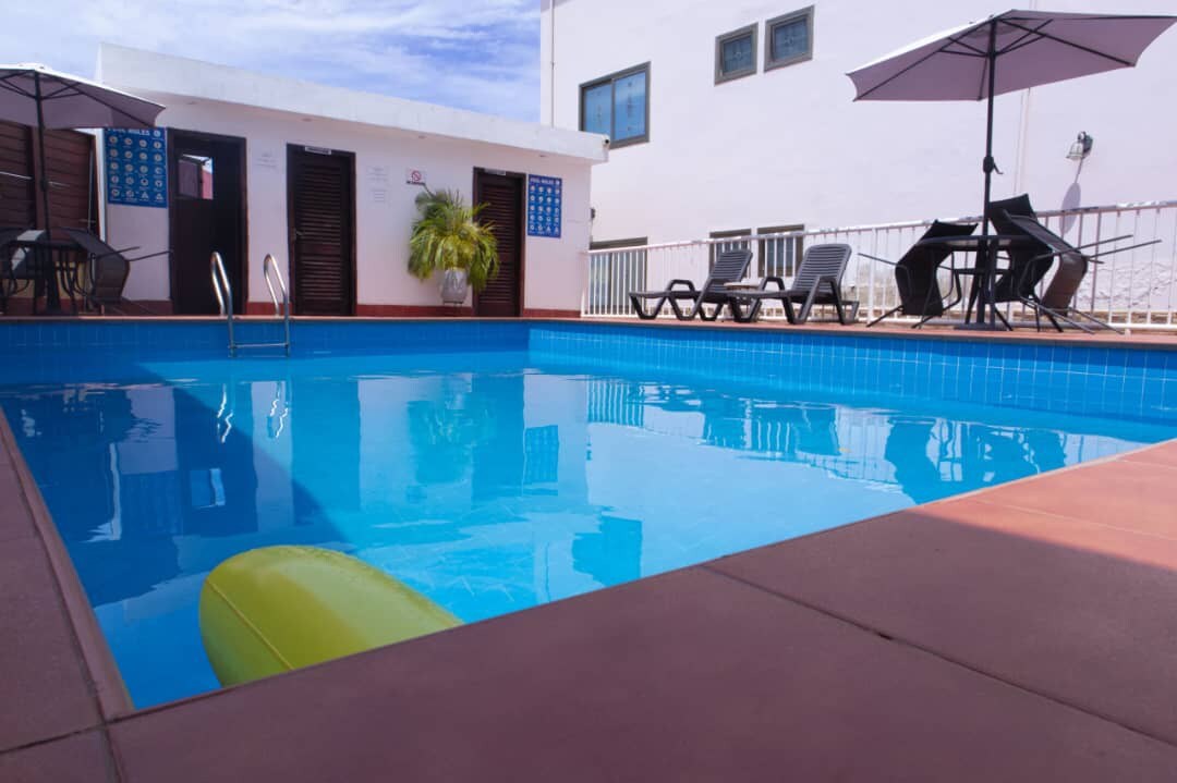 A refreshing blue swimming pool is surrounded by lounge chairs and shaded tables. Two umbrellas provide shade, while a pool ladder leads into the crystal-clear water. A bright yellow float is partly visible in the water, enhancing the tranquil setting.