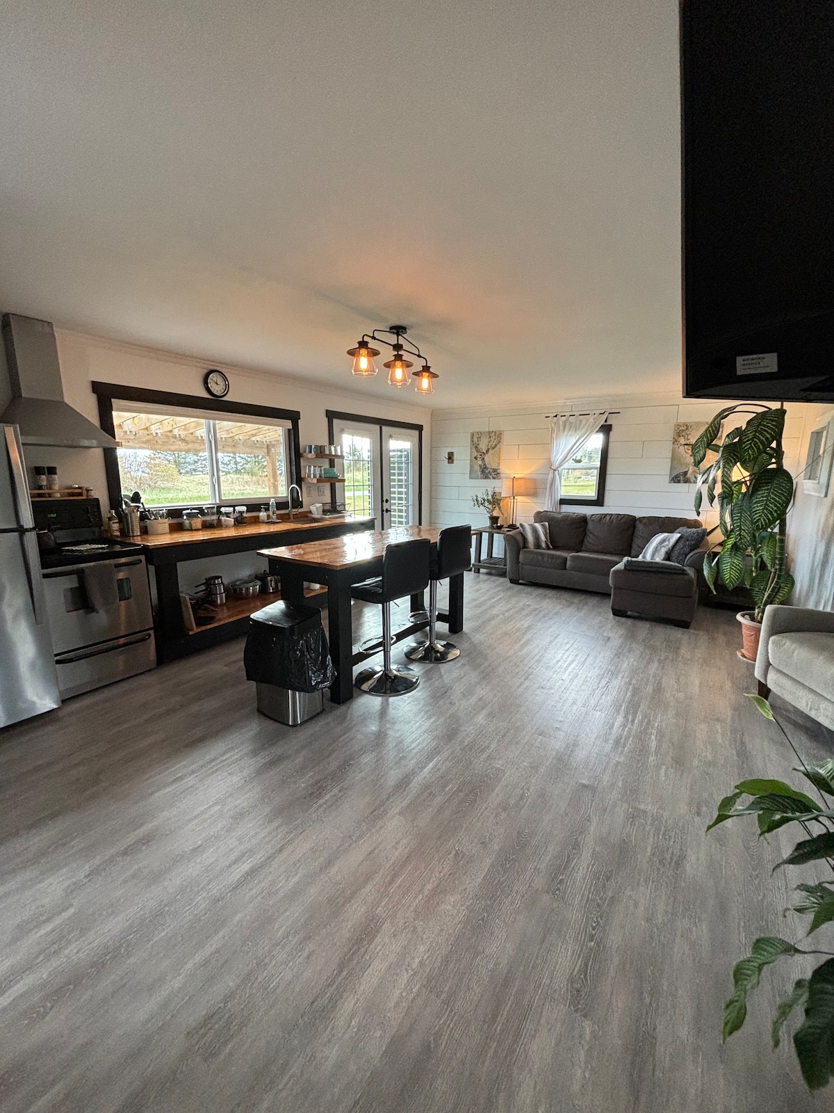 An open living space features a modern kitchen with stainless steel appliances and a large window providing natural light. A dining area with high stools and a cozy seating area is visible, complemented by greenery from nearby plants.
