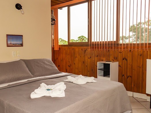 A cozy sleeping area features a spacious bed with smooth, neutral bedding. Freshly arranged towels are placed on the bed, and natural light filters in through large windows adorned with vertical blinds, offering an inviting view of the surroundings.