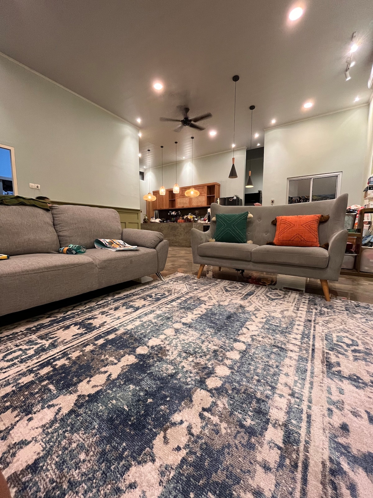 A spacious living area is furnished with a gray sofa and a mid-century style chair, both adorned with colorful throw pillows. A patterned area rug covers the floor, and modern pendant lights illuminate the room, highlighting the green walls and open layout that connects to the kitchen.