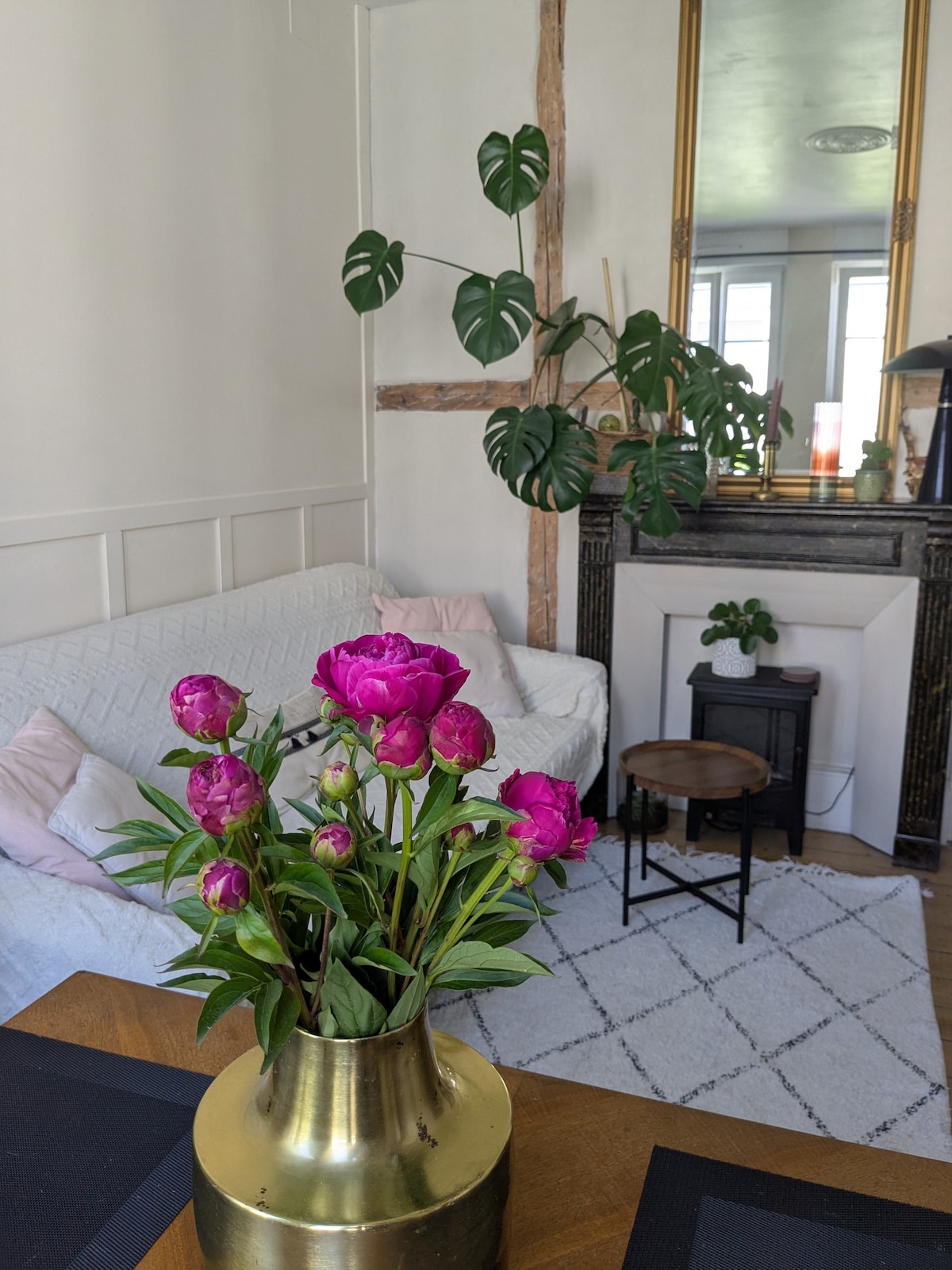 A spacious living area is showcased, featuring a white sofa adorned with soft cushions. A decorative fireplace is complemented by a tall mirror above it. A brass vase filled with vibrant pink peonies sits on a table, while a small plant rests beside a black side table.