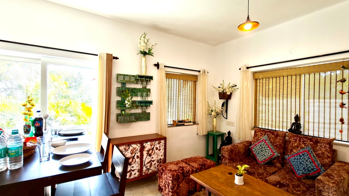 A welcoming living area features a comfortable sofa and patterned chairs, accompanied by a dining table set for guests. Natural light pours in through large windows, adorned with bamboo blinds, while decorative plants and artwork enhance the ambience.