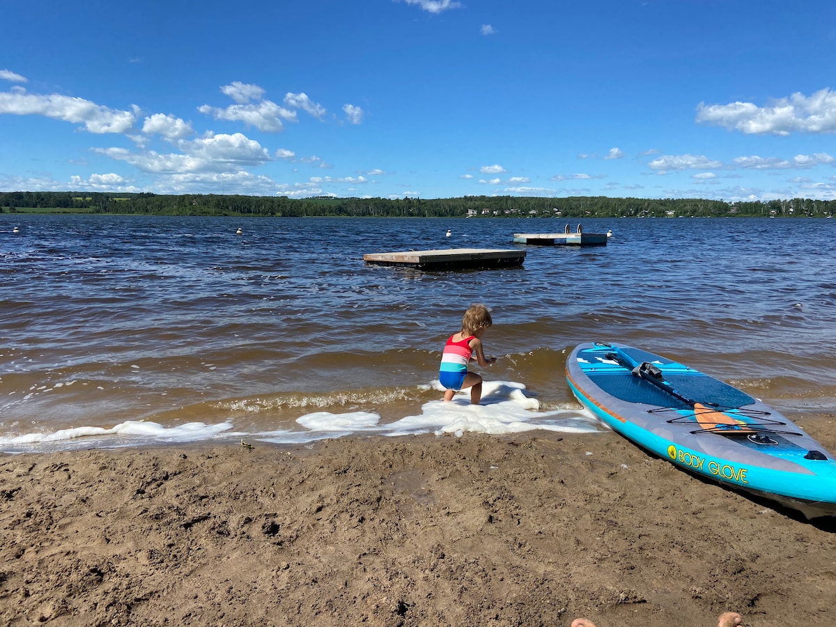 A sandy beach area is depicted, featuring gentle waves lapping at the shore. A child in a striped swimsuit plays at the water's edge. A paddleboard rests nearby, with calm water stretching toward distant floating docks under a blue sky with scattered clouds.