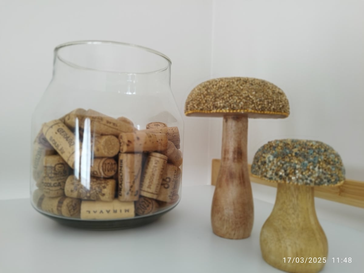 A glass jar filled with wine corks is displayed prominently. Next to it, two handmade mushroom figures feature wooden stems and textured caps, one with a golden finish and the other with a sparkling blue top. The arrangement adds a decorative touch.