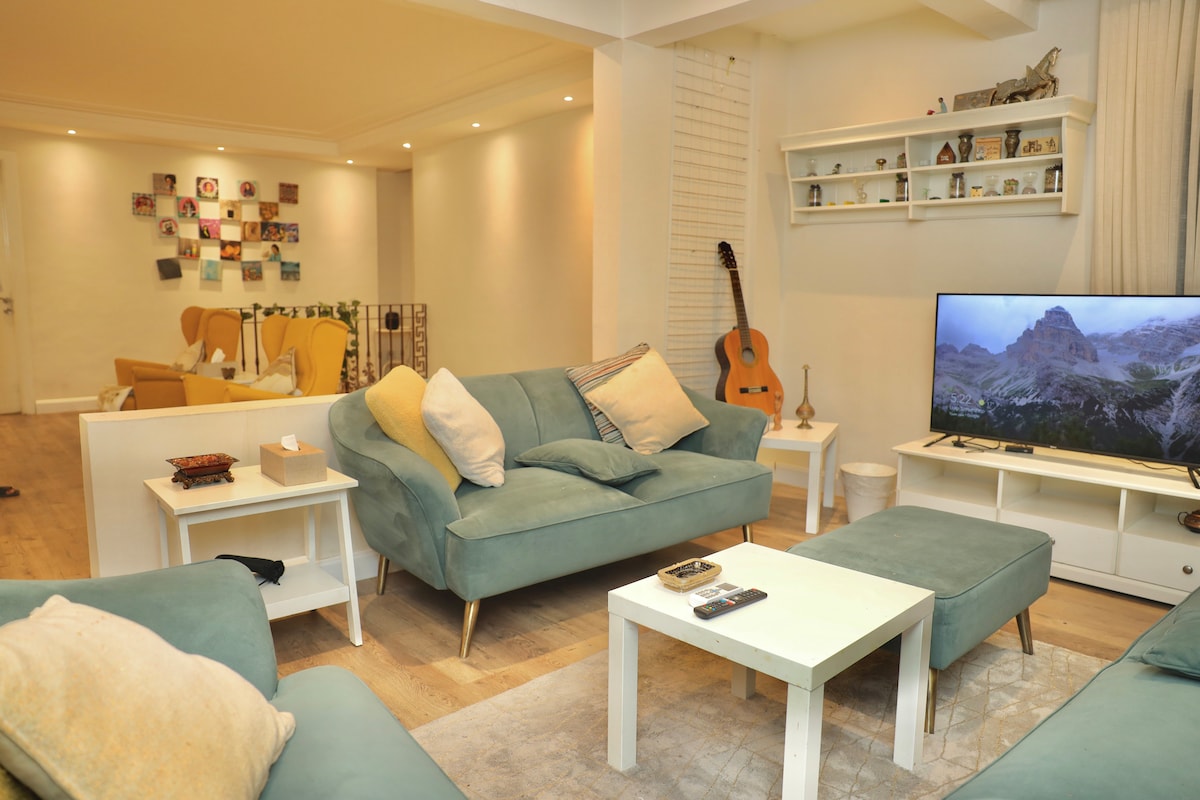 A comfortable living area features light green seating arrangements, with a white coffee table positioned centrally. A television is displayed against a light wall, while decorative shelves and framed photos create an inviting ambiance. Warm lighting enhances the spacious layout.