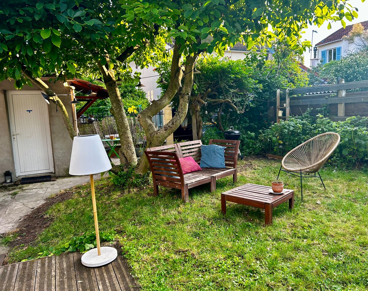 A private garden area is depicted, featuring a wooden seating arrangement with colorful cushions and a small coffee table. A modern floor lamp stands beside the seating, while a tree provides natural shade. The entrance to the mini house is visible in the background.
