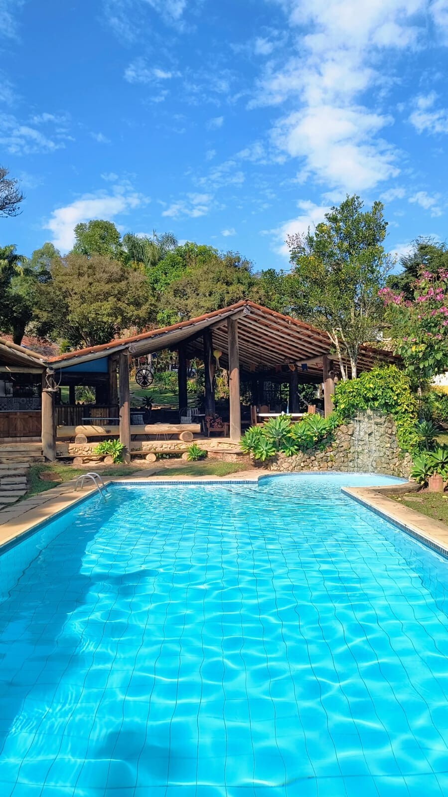 A clear blue swimming pool is framed by lush greenery. A rustic structure with a wooden roof and open design provides shaded seating areas. Trees and flowering plants enhance the natural surroundings, creating a relaxing outdoor environment.