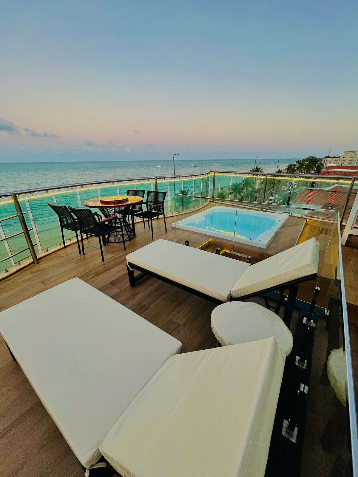 An outdoor terrace overlooks the sea, featuring a private Jacuzzi and a comfortable seating area with lounge chairs. A dining table with chairs is positioned nearby, and the expansive view captures the horizon against a pastel sky during sunset.