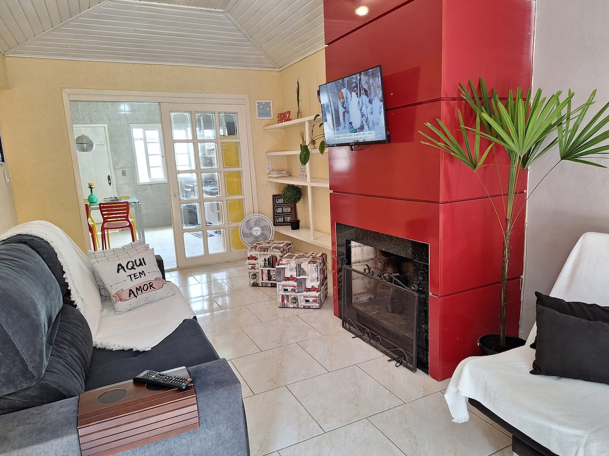 A cozy living area is featured, highlighted by a red accent wall and a comfortable seating arrangement with a three-seat sofa and armchair. A television is mounted on the wall, and a wall fan is positioned nearby. A fireplace adds a warm focal point, enhancing the inviting environment.