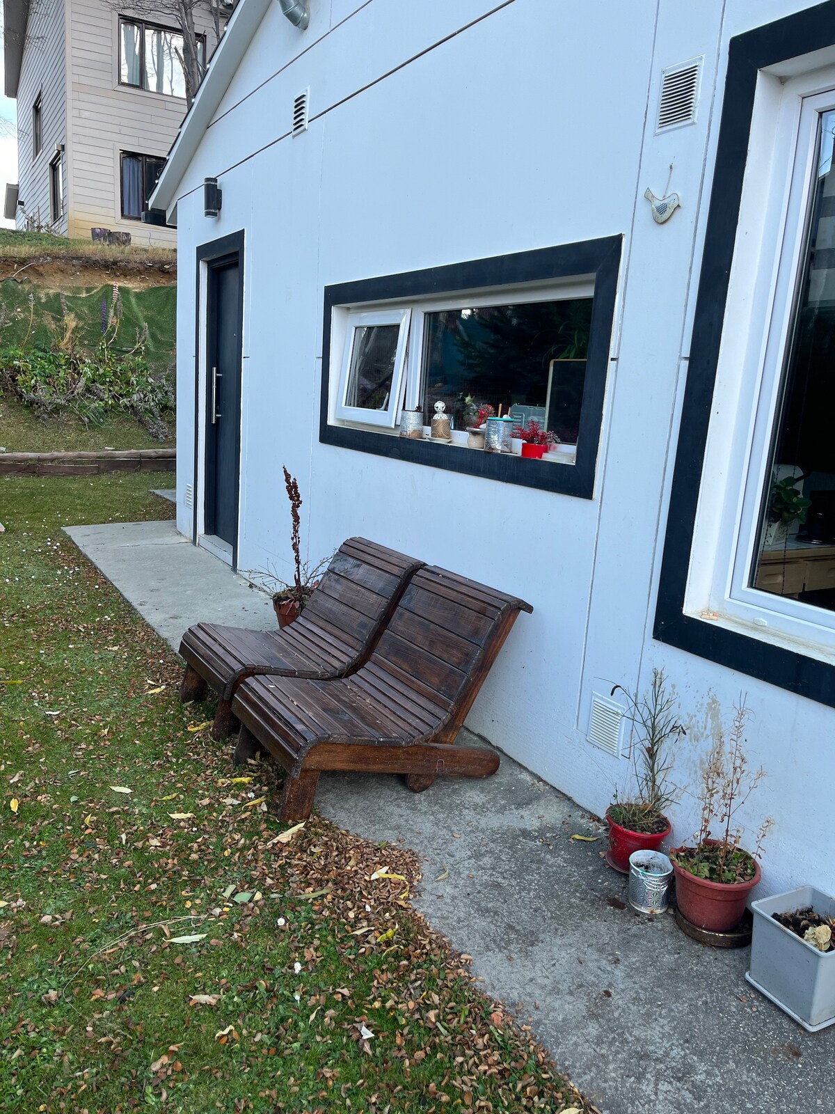 A wooden bench is positioned against the exterior wall of the house, offering a space to relax. Small potted plants are placed along the ground, adding a touch of greenery. A window with a view of the interior is visible, allowing natural light to enter.
