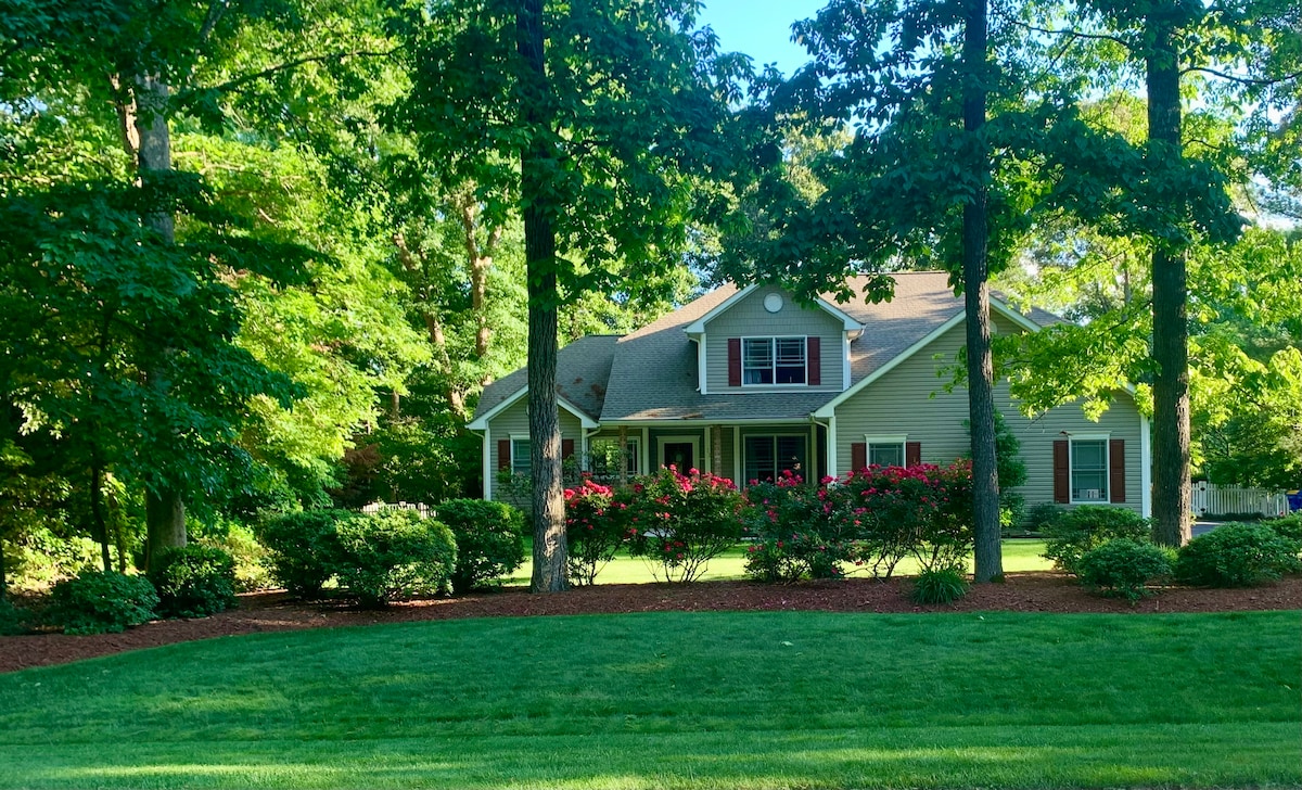 A charming home is surrounded by lush greenery and vibrant flowering shrubs, set against a clear, blue sky. The inviting facade features large windows and a welcoming front porch, offering a glimpse into a serene residential environment.
