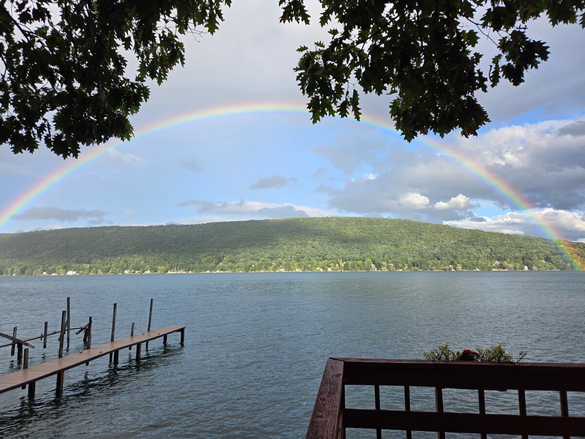 A rainbow arcs across the sky above a calm lake, with a lush green hillside in the background. The image captures a view from a wooden deck overlooking the water, framed by overhanging tree branches.