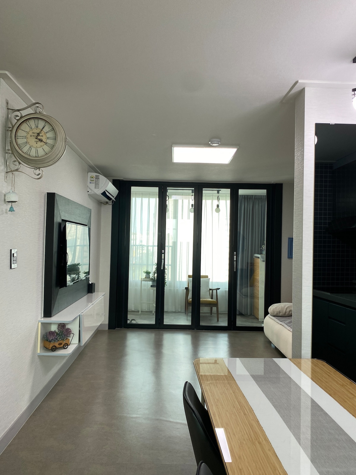 A modern living space is highlighted by a large wall-mounted television and a stylish table with chairs. Floor-to-ceiling glass doors allow natural light, leading to a balcony area. Neutral-toned walls and minimalistic decor create a clean and open atmosphere.