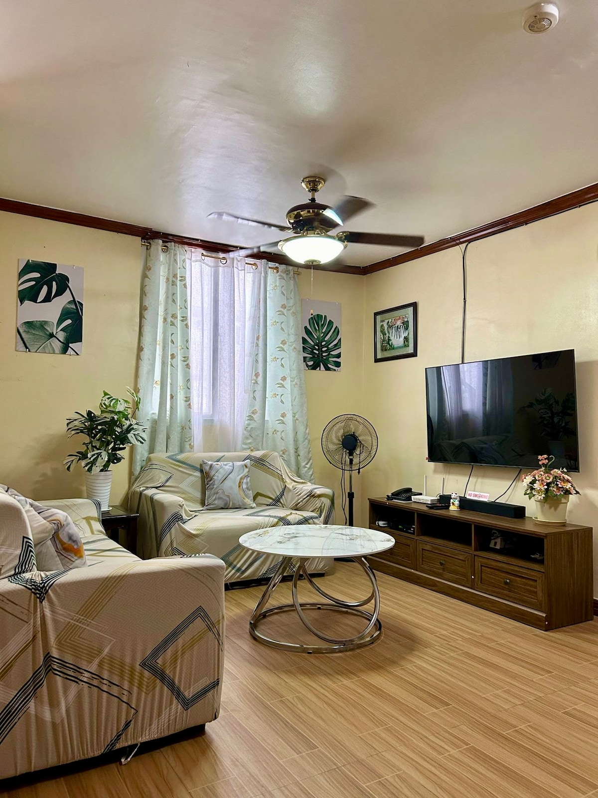A spacious living room features a comfortable seating area with light-colored furniture and decorative cushions. A round coffee table occupies the center, while a large wall-mounted TV is positioned on a wooden media unit. Natural light filters through partially draped windows, enhancing the cozy ambiance.