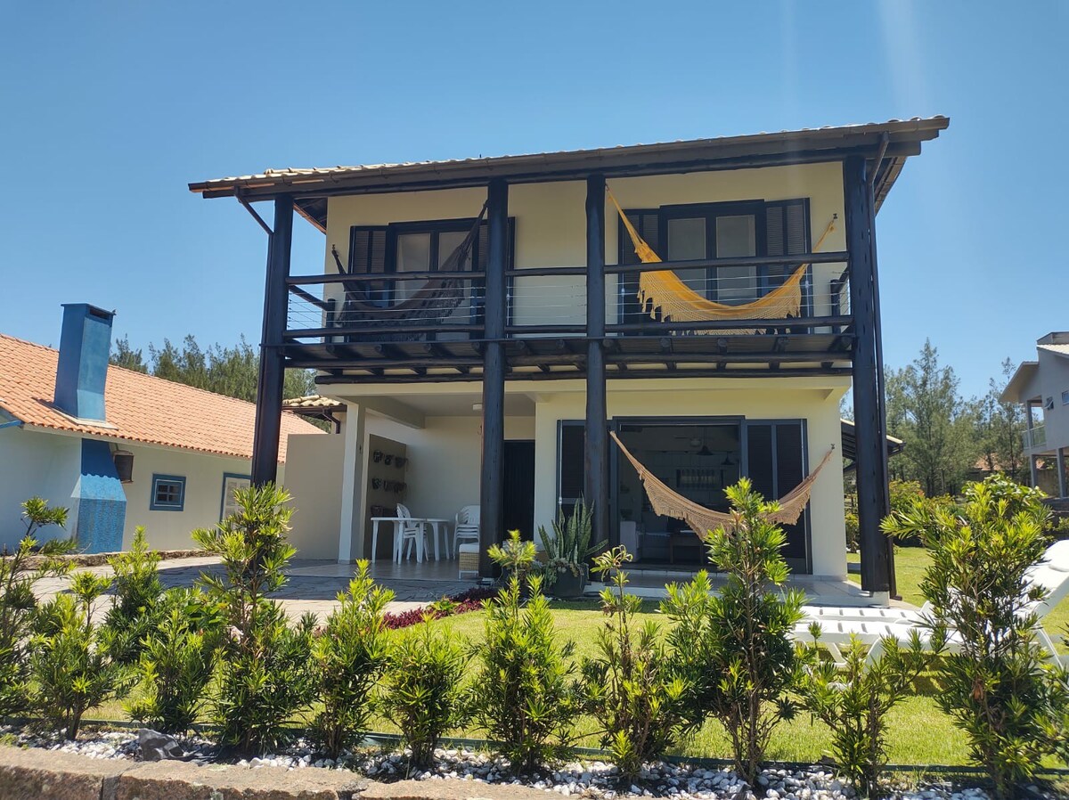 The two-story house features a classic design with a brown wooden frame and large windows. Hammocks are draped on the balconies, while a manicured lawn and landscaped plants surround the front area, providing a welcoming outdoor space.