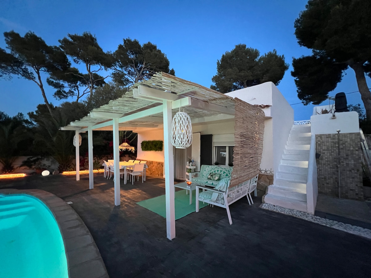 A charming outdoor seating area features a covered terrace with a dining table and chairs, surrounded by lush greenery. Soft lighting illuminates the space, while a staircase leads to the upper level of the villa. The inviting pool reflects the evening sky.