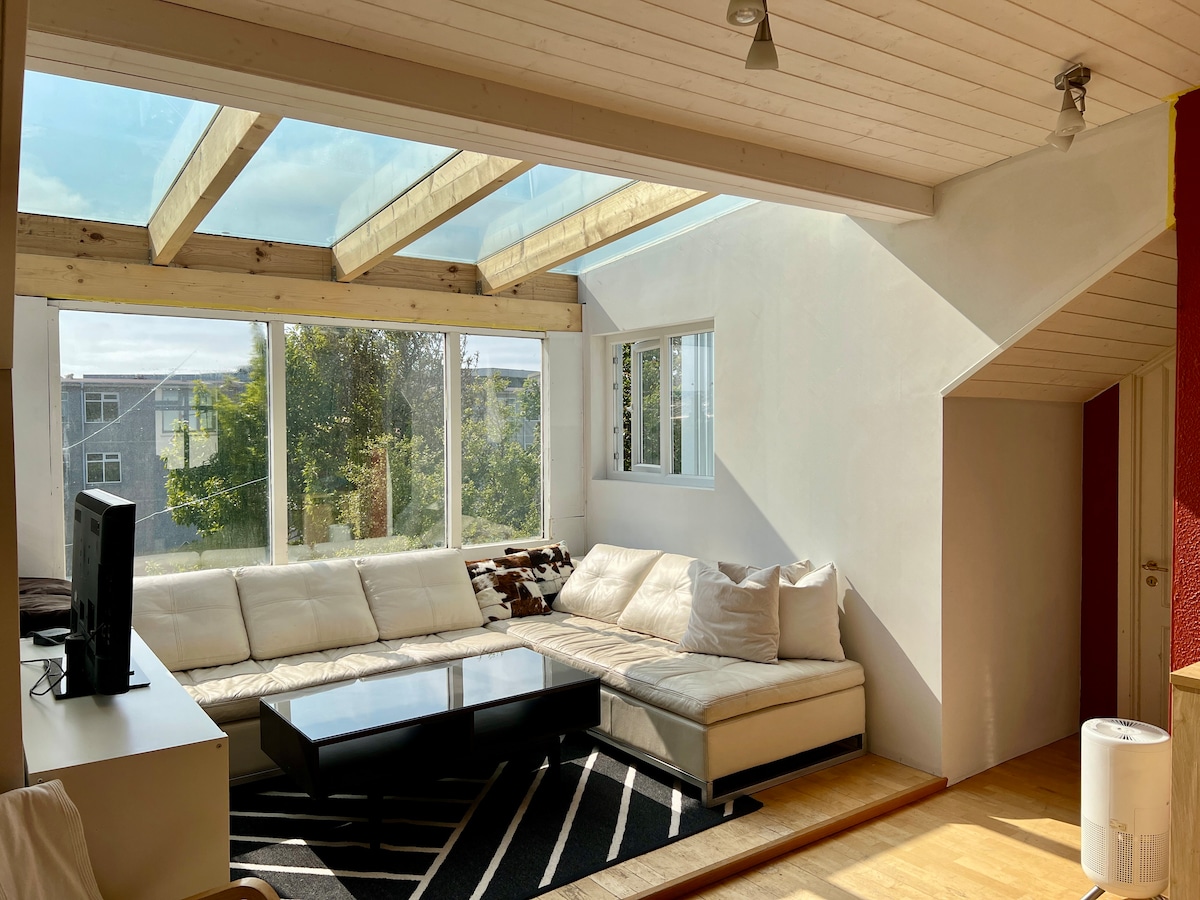 A light-filled living area features a spacious sectional sofa and a rectangular coffee table on a patterned rug. Skylights and large windows provide ample natural light, while greenery is visible outside, enhancing the sense of openness.